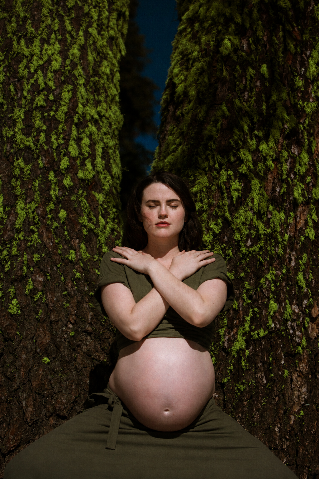 A woman in an olive dress sitting peacefully between two moss-covered trees